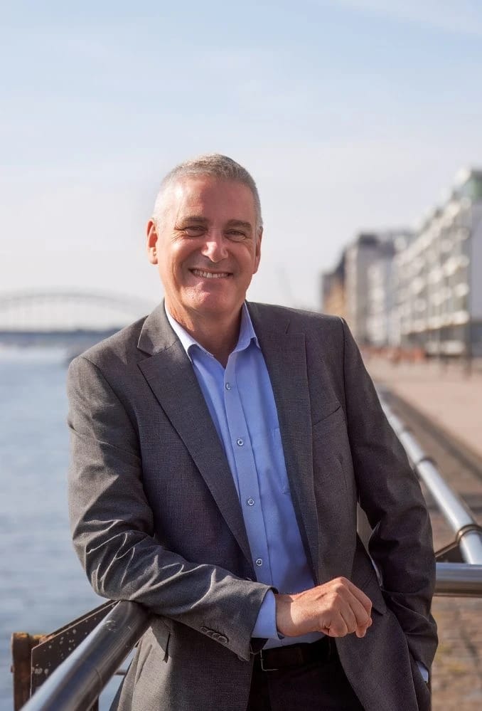 Bernd Schäfer im grauen Sakko lächelt an einer Uferpromenade in die Kamera