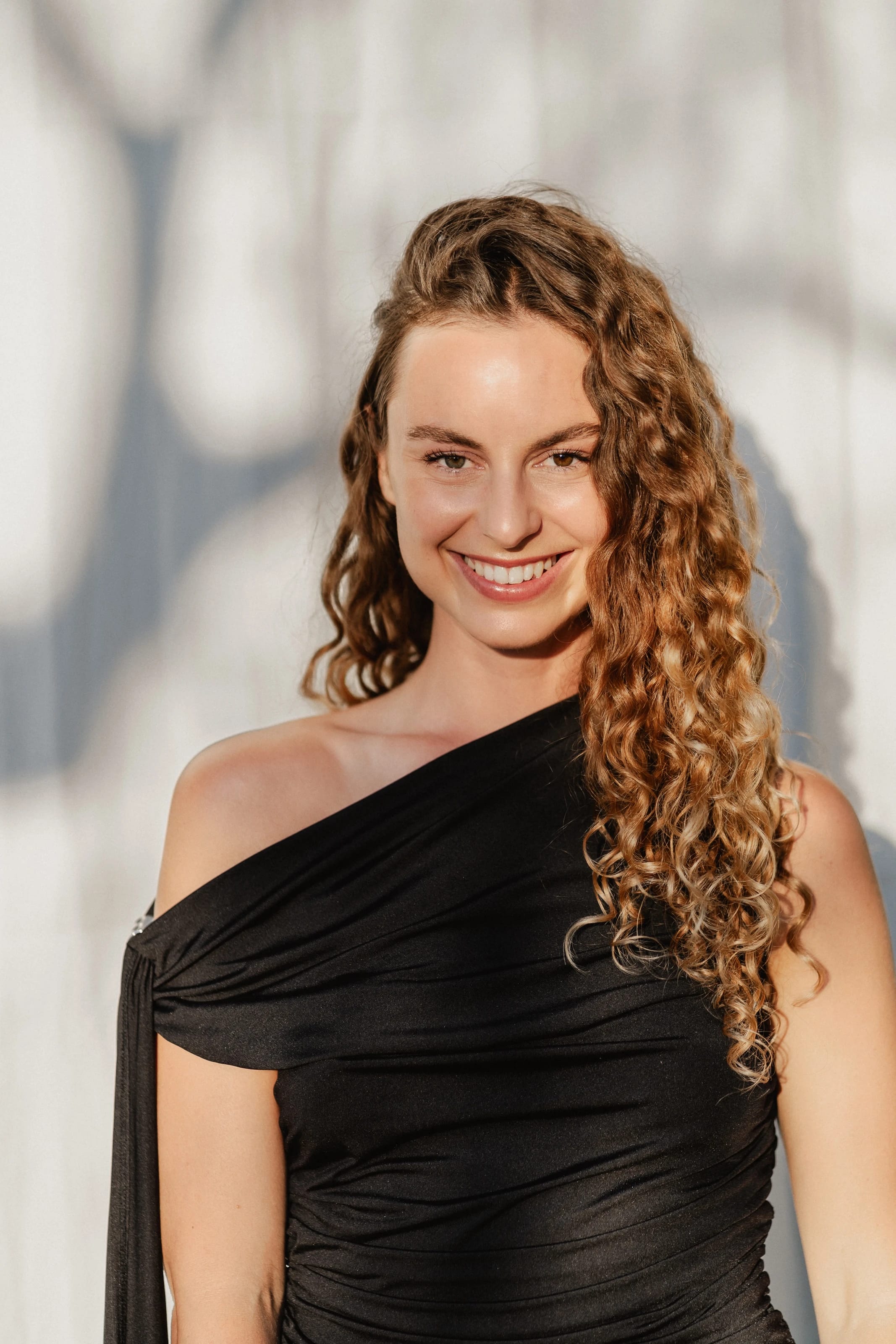 Hannah lächelt in schwarzem One-Shoulder-Kleid vor heller Wand