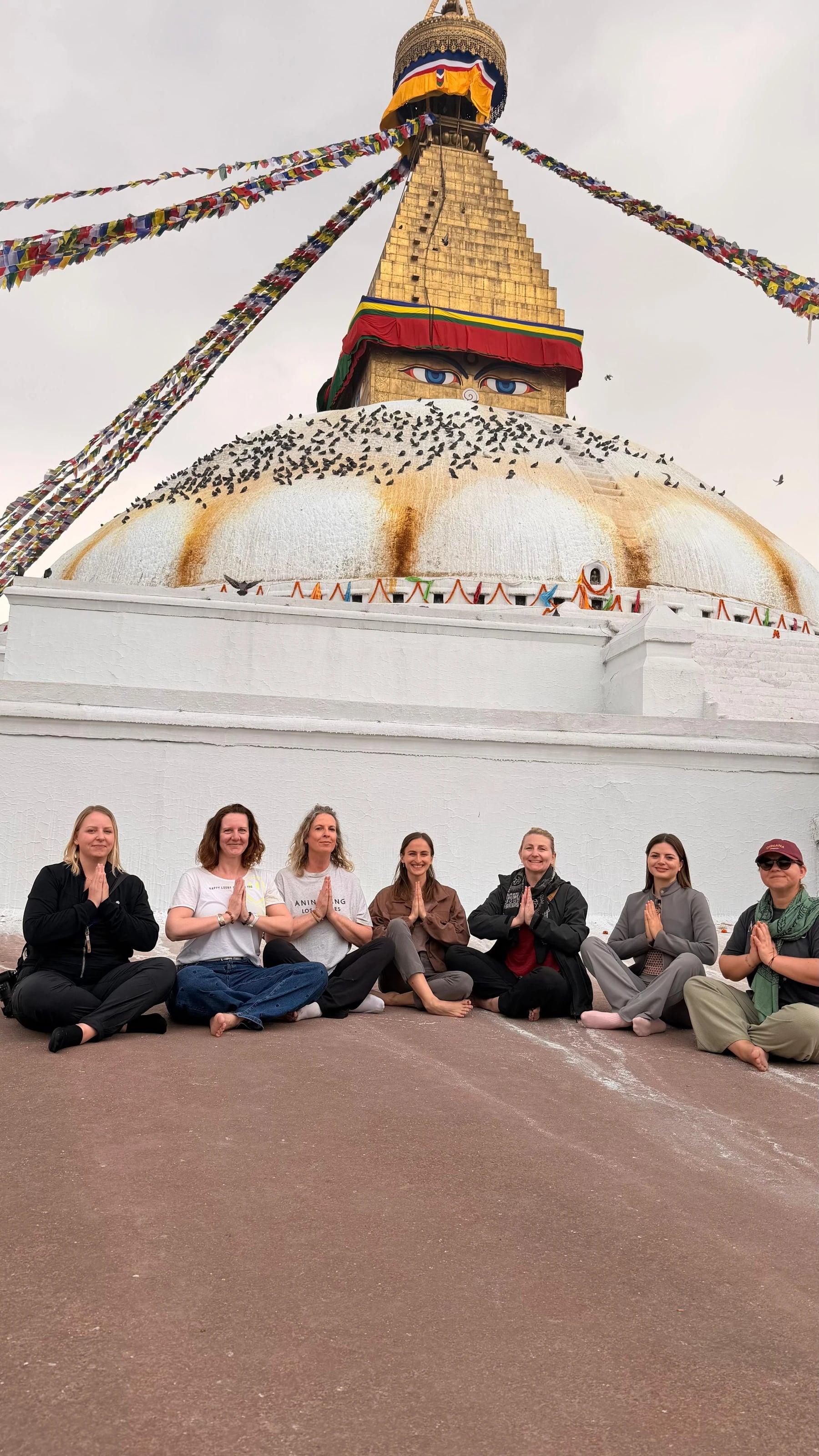 Reisegruppe sitzt mit gefalteten Händen vor der tauben-umschwärmten Stupa von Boudhanath.
