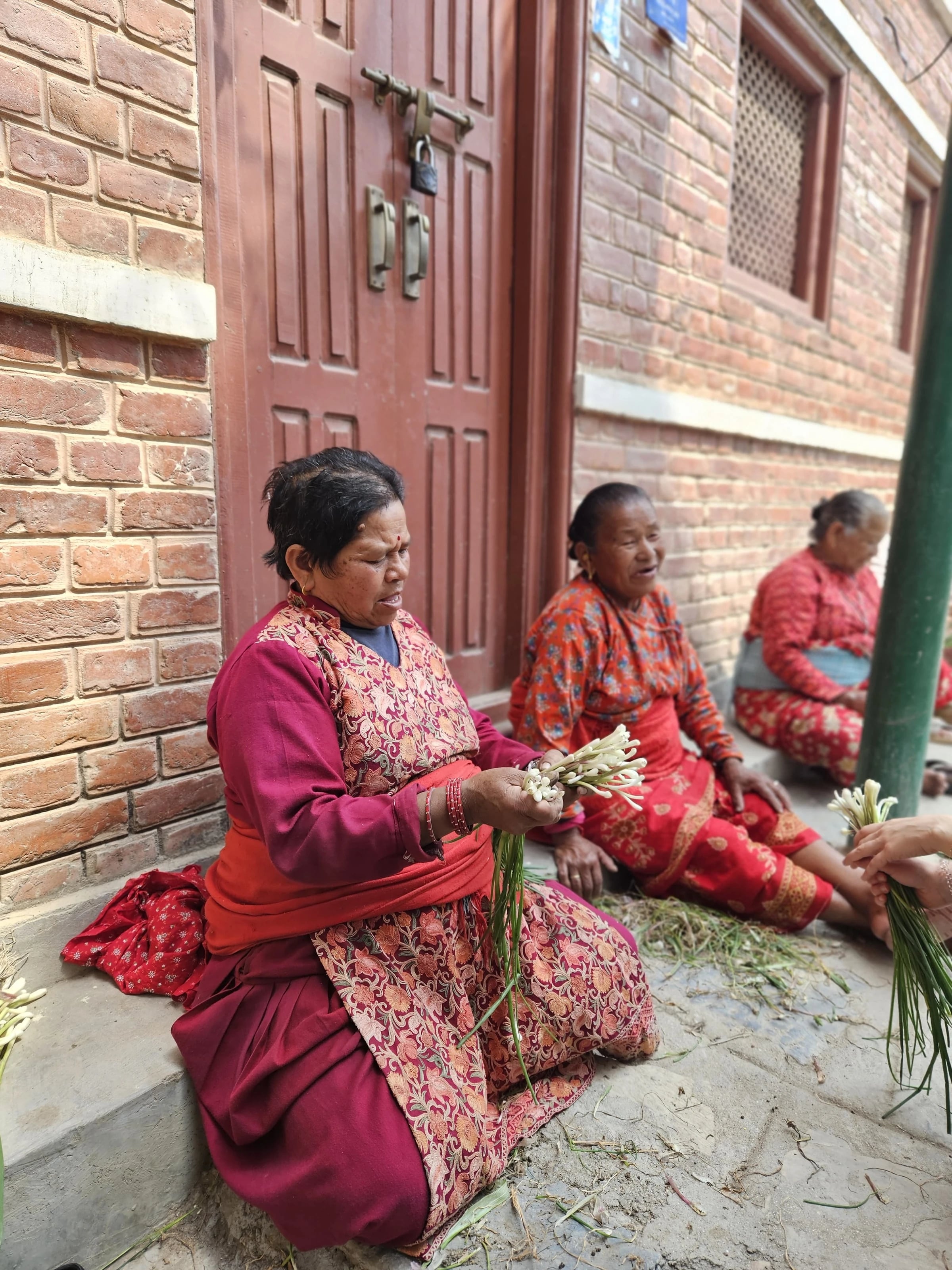 Einheimische Frauen fädeln Blumen vor einer roten Backsteinhaustür in Nepal.