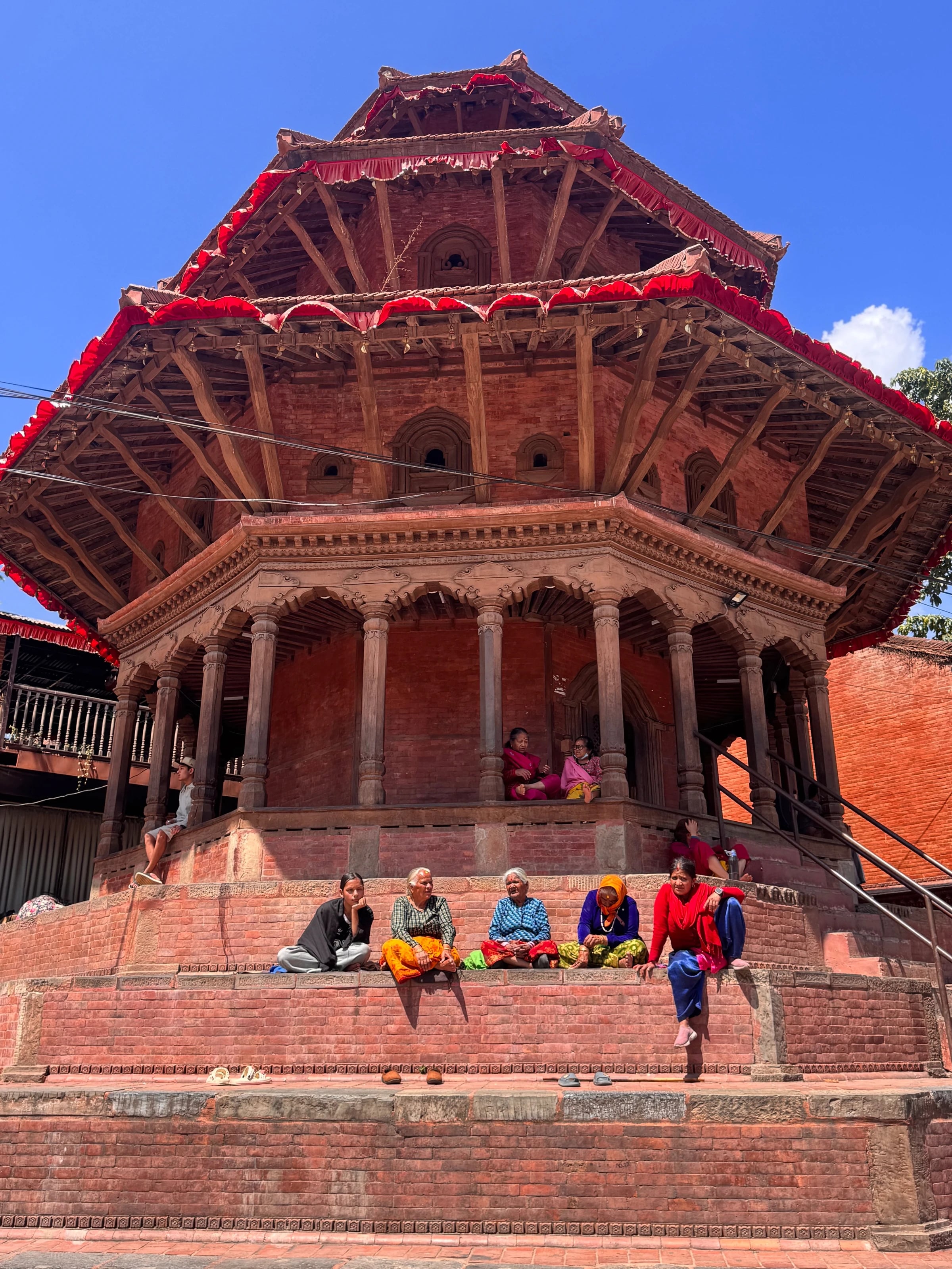 Ein rot-goldener Tempel am Durbar Square in Patan, davor sitzen einheimische Frauen auf den Stufen.