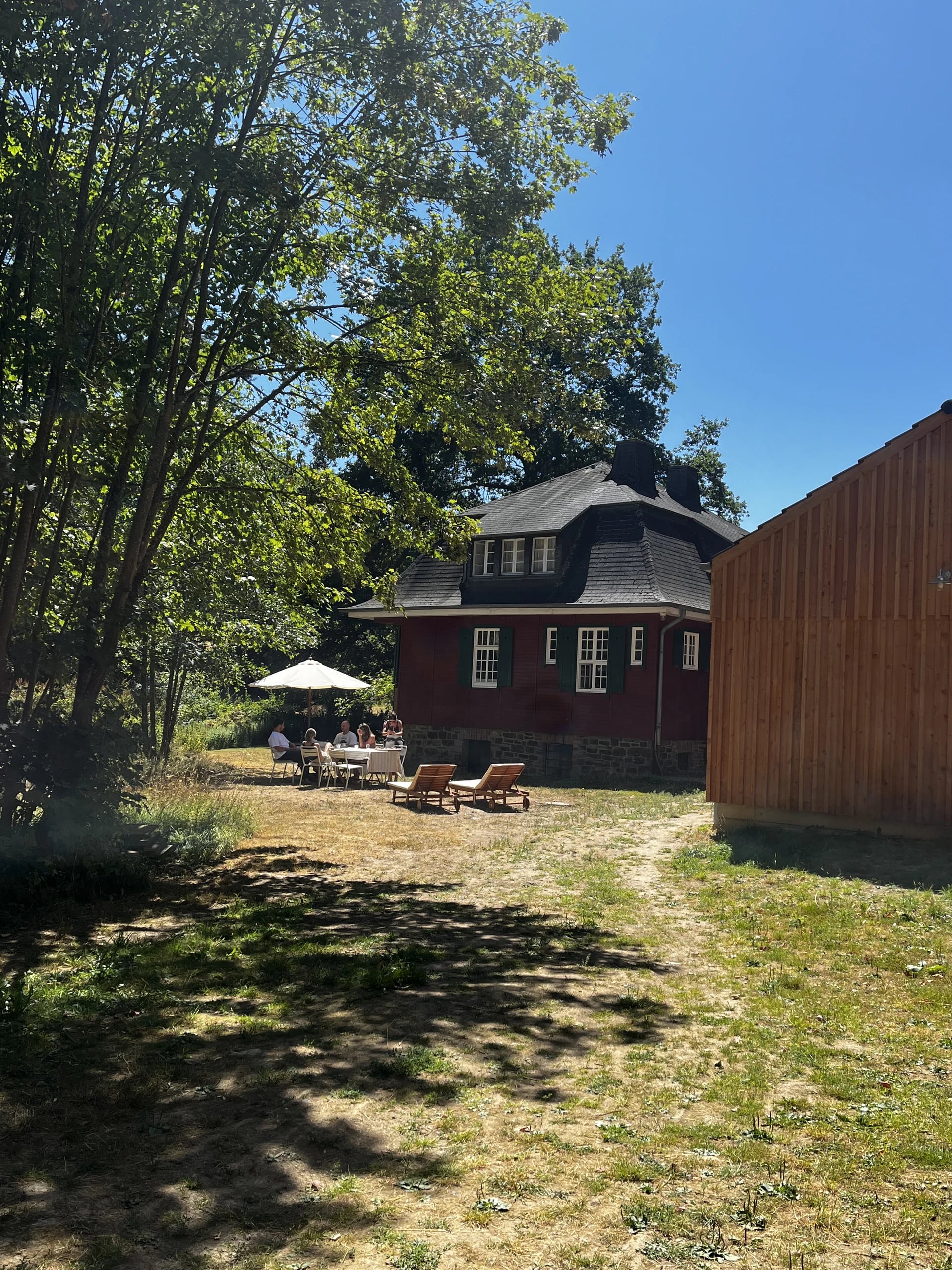 Das rote Haupthaus mit Schieferdach, davor sitzen Frauen an einem gedeckten Tisch unter einem Sonnenschirm.