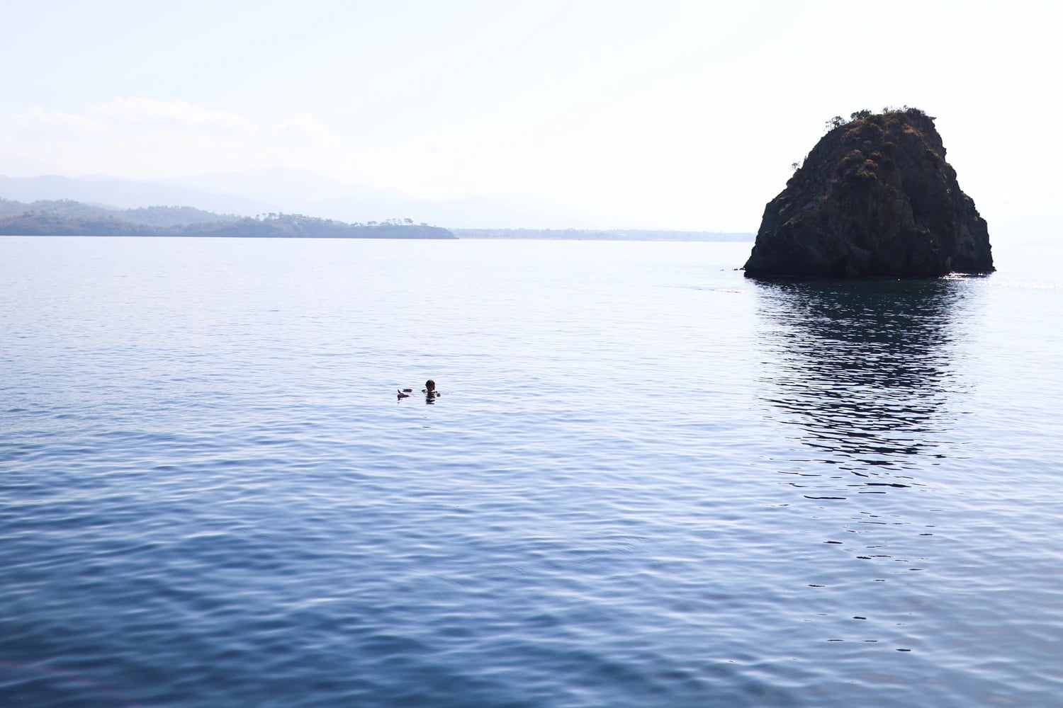 Eine Schwimmerin im weiten, ruhigen Meer neben einem markanten Felsen