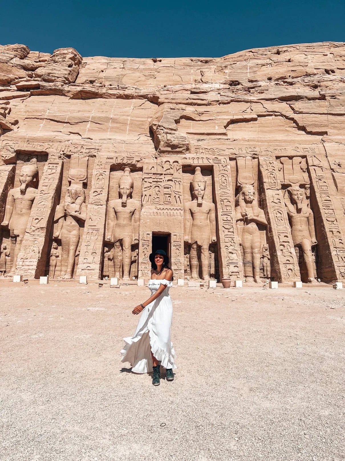 Eine Frau in weißem, wehendem Kleid und Hut steht vor den kolossalen Felsstatuen der Tempelfassade von Abu Simbel.