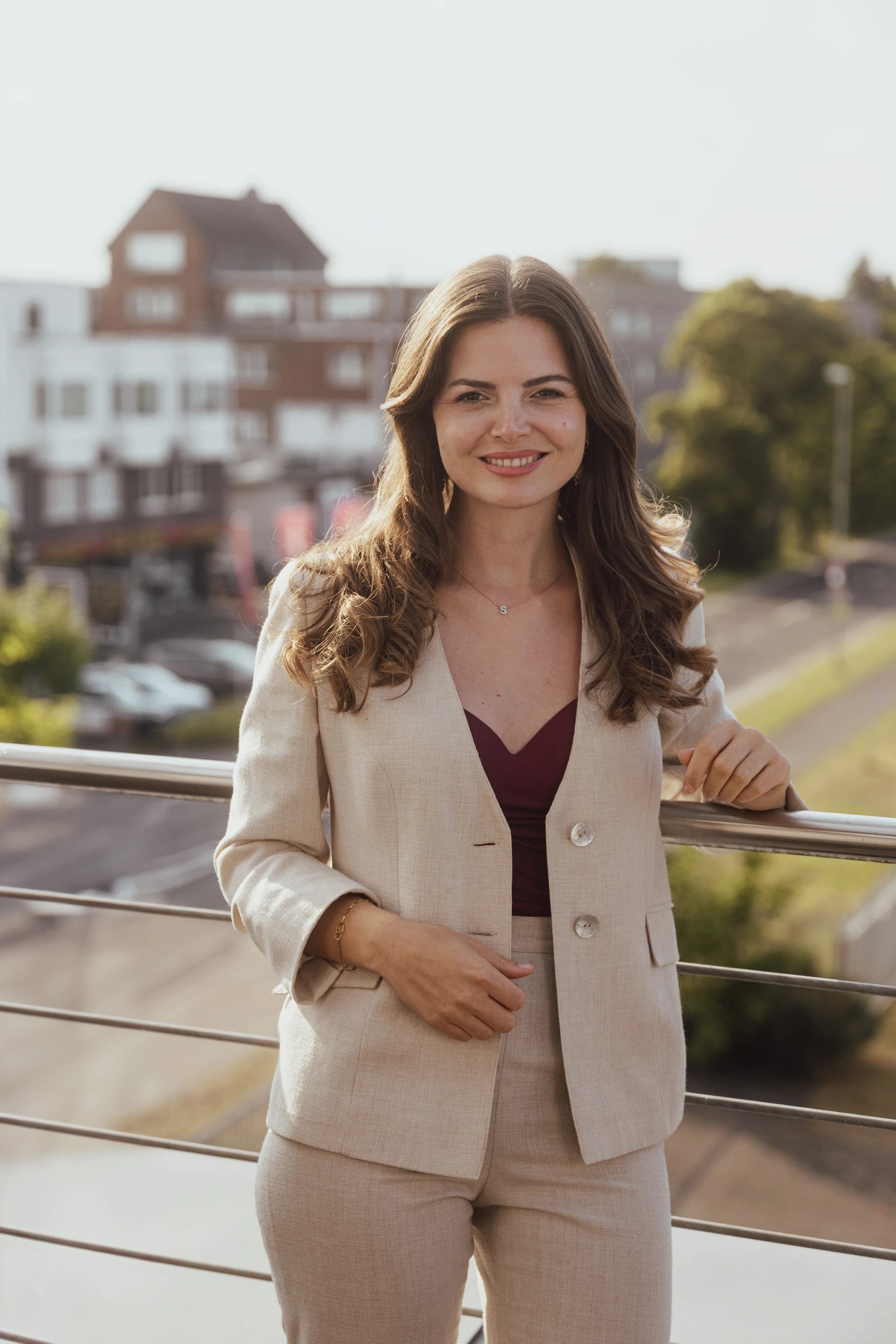 Selin im beigen Anzug, lächelnd, vor einer europäischen Stadtkulisse