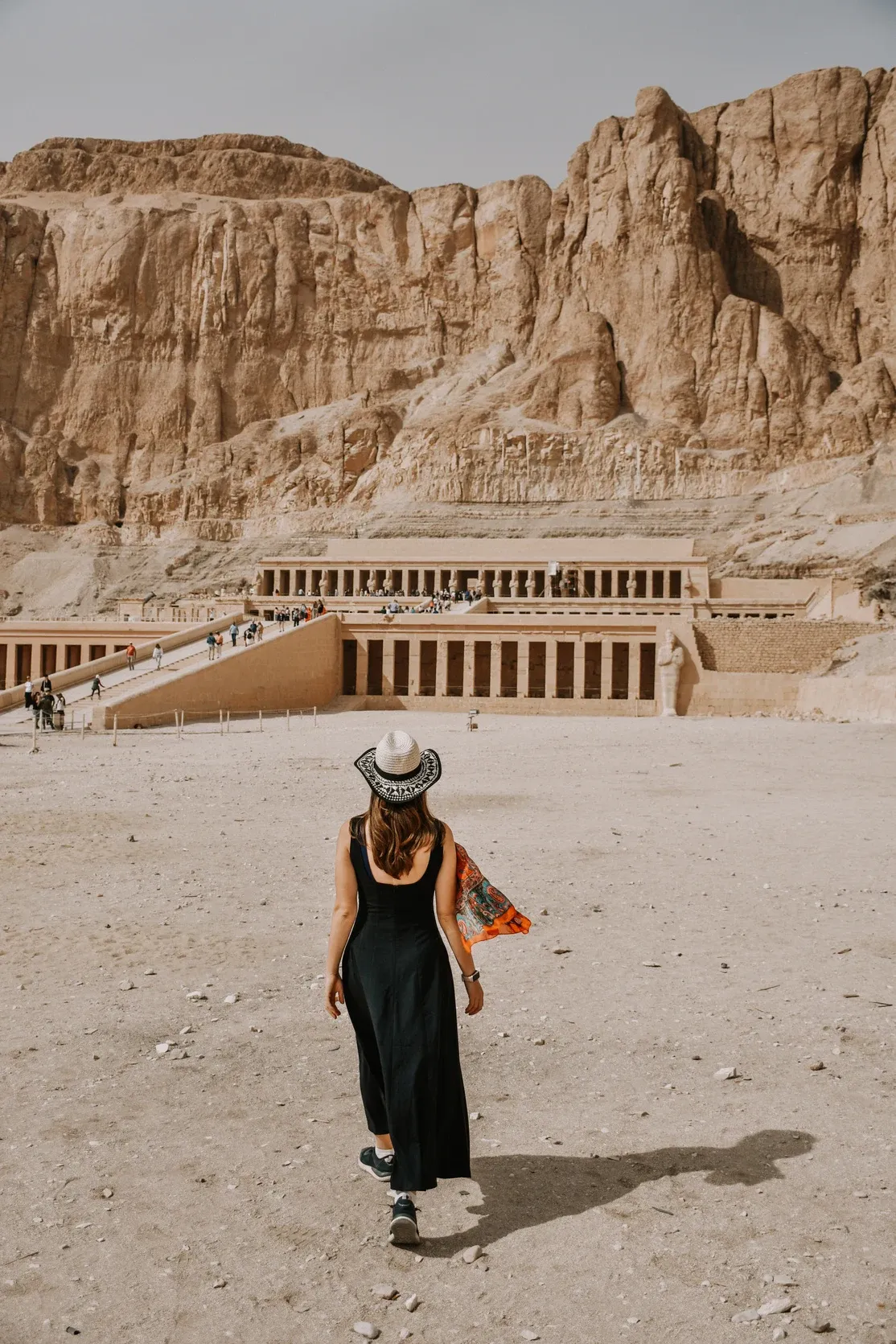 Eine Frau in schwarzem Kleid und Hut geht von hinten gesehen auf den Hatschepsut-Tempel unter hohen Felsklippen zu.