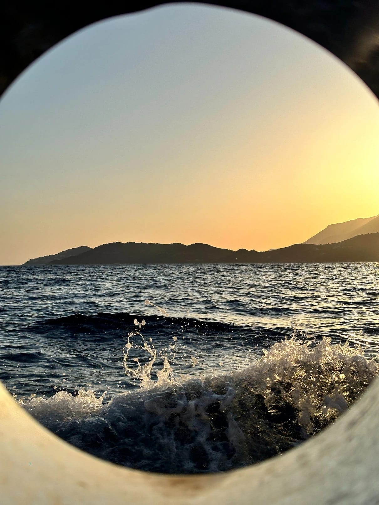 Blick durch ein rundes Bullauge auf eine brechende Welle und den Sonnenuntergang über der türkischen Küste