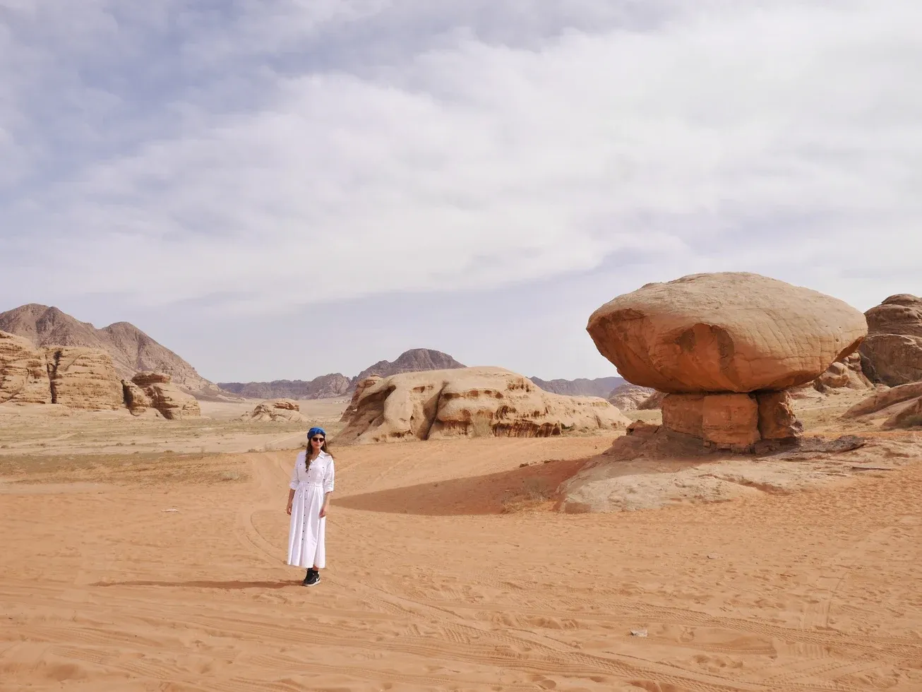 Eine Frau in weißem Kleid steht in der Wüste neben einer großen pilzförmigen Felsformation in Wadi Rum.