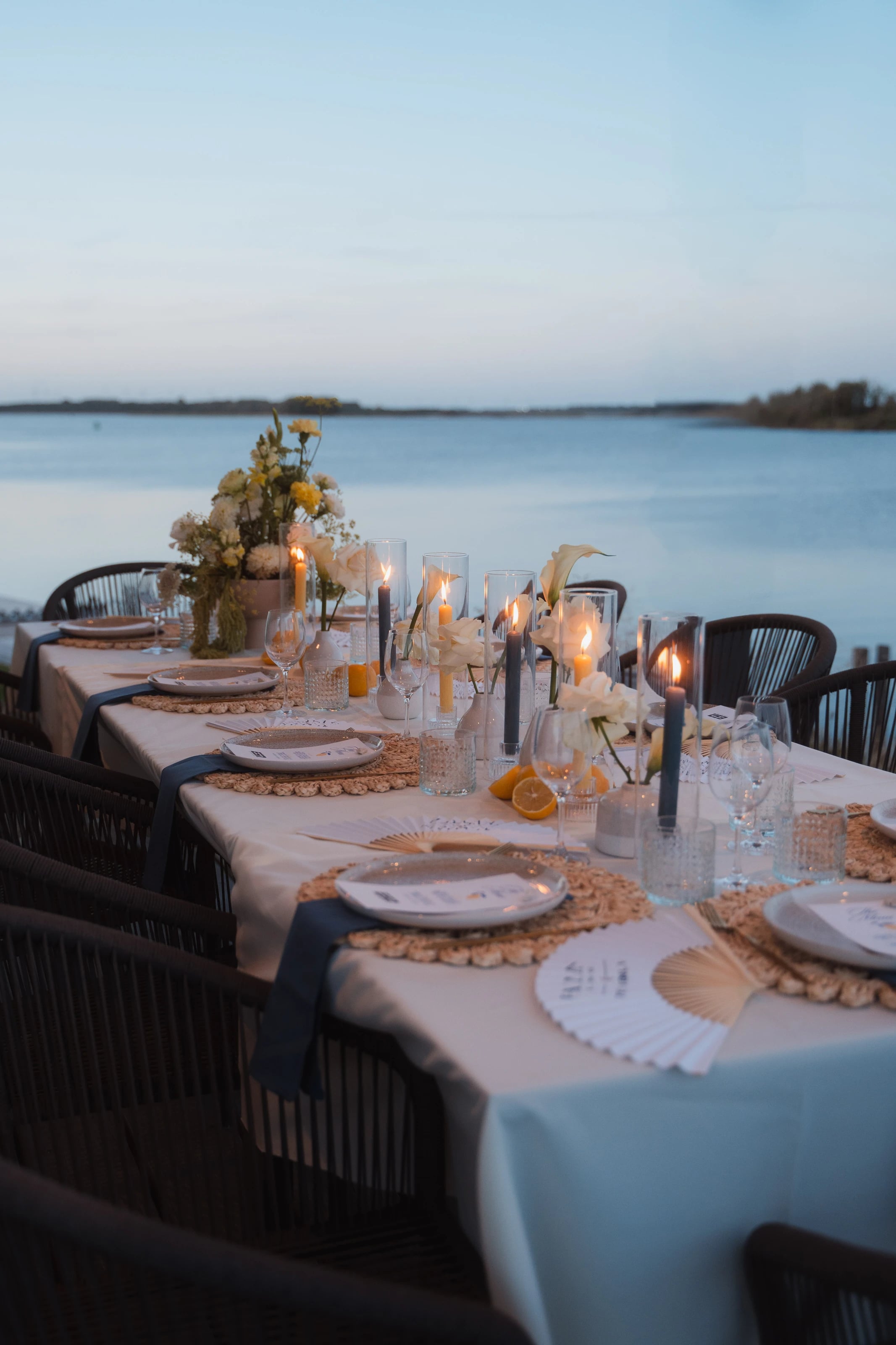 Eine lange Tafel am Wasser mit Kerzen und gelben Blumen zur blauen Stunde.